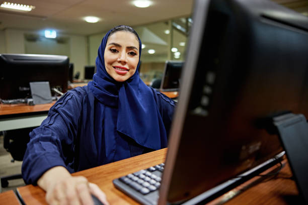 Portrait of smiling Saudi woman in abaya sitting at desk in computer lab